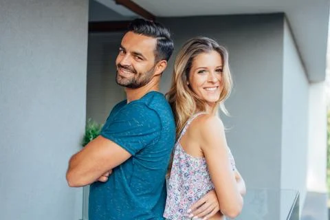 Young couple is standing back to back at the balcony and smiling. City backgr Stock Photos