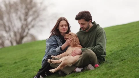 Young couple talking and playing with dog. Dog owners sitting on the grass Stock Footage 240290265