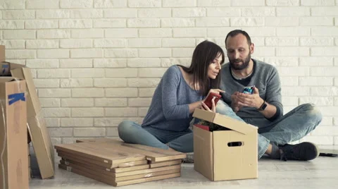 Young couple talking and using smartphone while sitting on floor at new home Stock Footage 65382886