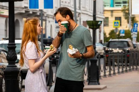Young couple talking while walking along city street and eating takeaway food Stock Photos