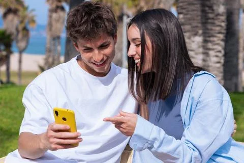 Young couple using smartphone on bench under palm trees smiling at phone social Stock Photos