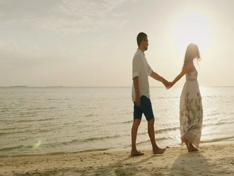 A young couple is walking along the beach at sunset. A woman leads a man. Follow Stock Footage 80081072