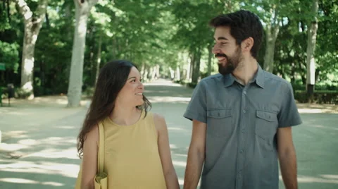 Young couple walking hand to hand in the park, talking and laughing. Dolly shot. Stock Footage 64745873
