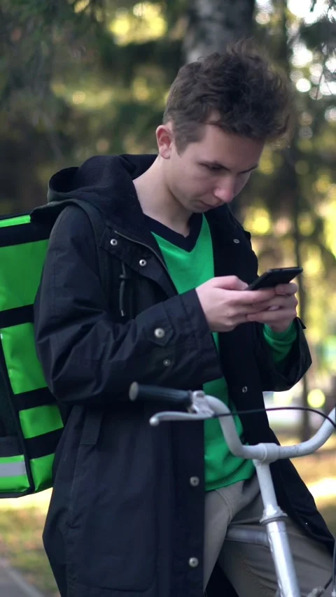 Young courier with green backpack checks his phone beside a bicycle in an autumn Video stock 346859507