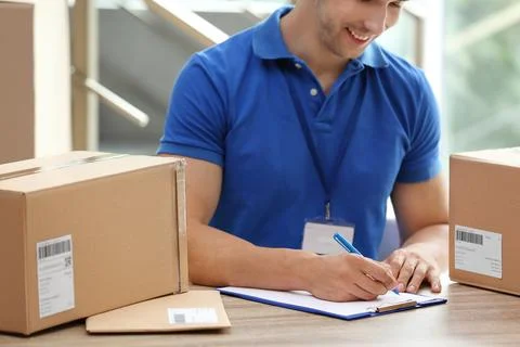 Young courier working with papers among parcels at table in delivery departme Fotos Stock