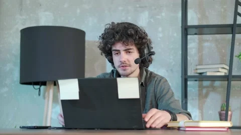 Young curly haired man using laptop computer with headset to talking online. Stock Footage 155759890