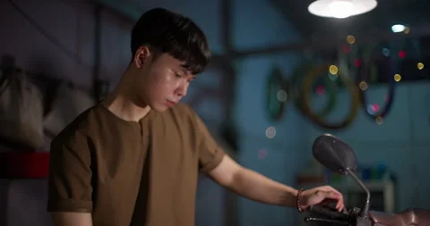 Young Customer Working Beside Moped Under Lamp in Dim Repair Shop Stock Footage 346410834