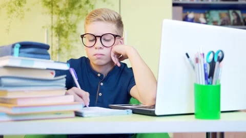 Young cute boy, schoolboy with glasses sitting at the table teaches lessons.  Stock Footage 138451516