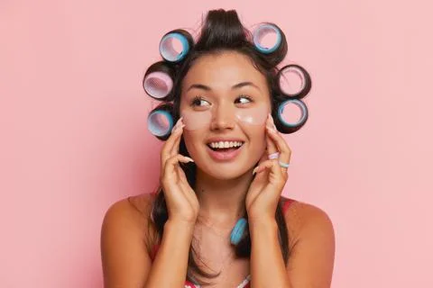 Young cute woman posing on pink backdrop, with curlers on black hair, pressing Stock-Fotos