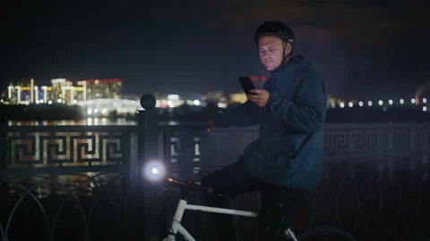 Young Cyclist Using Smartphone by River at Night with City Lights 库存影片 345708341