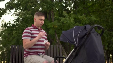 A young dad in the park on a bench with wireless headphones shakes a stroller Video stock 209353332