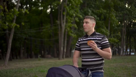 A young dad walks through the park with a stroller and eats a burger with a Video stock 206536835