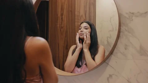 A young dark-skinned brunette in front of a mirror massages her face 스톡 동영상 162497428