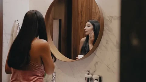 A young dark-skinned brunette in front of a mirror applies cream to her face. Video stock 162498288