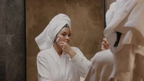 Young dark-skinned brunette in front of a mirror in a robe massages her face Video stock 162498871