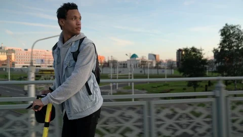 Young dark skinned guy with curly hair moves on a electric scooter on a bridge Stock Footage 115993018