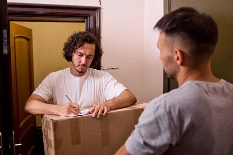 Young delivery worker handing large cardboard box to male recipient beside Stock Photos