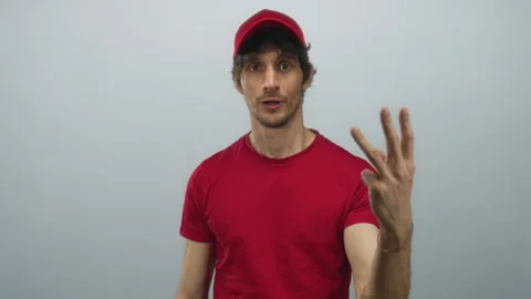 Young deliveryman in red cap shows two fingers at camera in grey studio; co.. Video stock 314298074