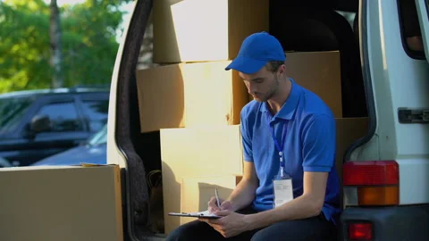 Young deliveryman writing parcel distribution report and smiling, sitting in van Stock Footage 91242899