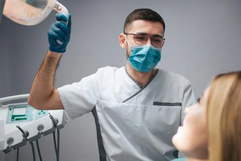 Young dentist in mask sit at client and hold lamp. Young woman look at doctor Stock Photos