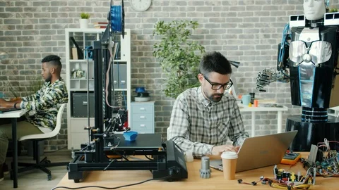Young developer using laptop typing in office, 3d printer and robot are visible Stock Footage 119581026