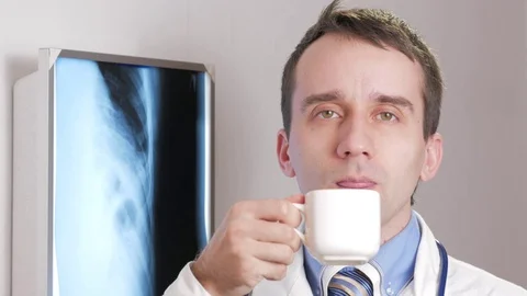 A young doctor looking at the camera is drinking a hot drink from a white mug Video stock 73956466