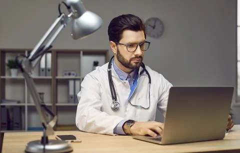 Young doctor working on a laptop computer while sitting at his desk at the Stock Photos