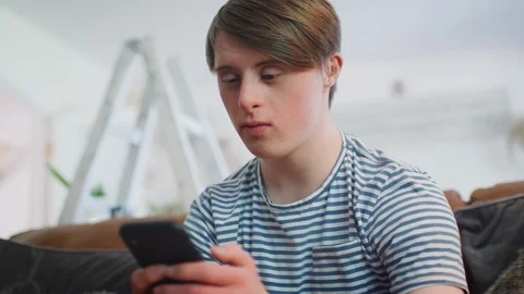 Young Downs Syndrome Man Sitting On Sofa Using Mobile Phone At Home Stock Footage 114305077