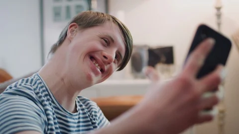 Young Downs Syndrome Man Sitting On Sofa Using Mobile Phone To Take Selfie At Stock Footage 114305085