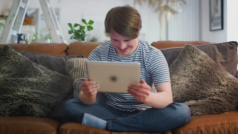 Young Downs Syndrome Man Sitting On Sofa Watching Digital Tablet At Home Stock Footage 114305143