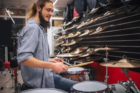 Young earded hipster sit close to drums and cumbals and playing on them. He hold Foto stock