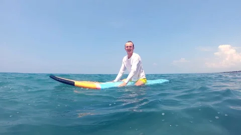 Young european-looking guy dressed in white blouse sitting on a surfboard Stock Footage 128959079