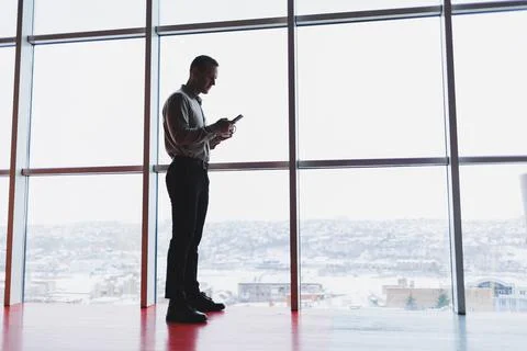 A young executive is holding a phone while standing in an office interior and Stock Photos
