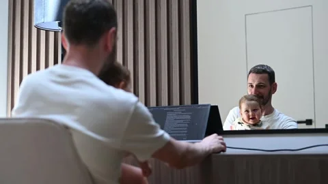 Young father and programmer working remotely from home on a laptop, holding his Stock Footage 323879559