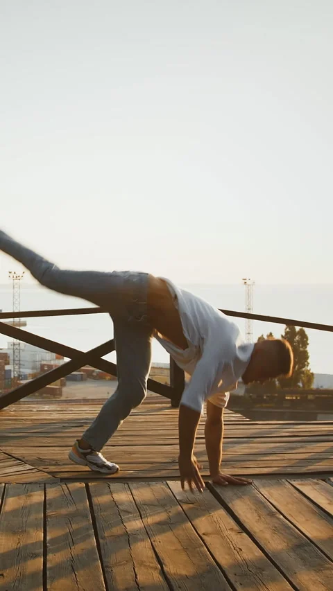 Young fellow is performing break dance on wooden terrace against picturesque Stock Footage 314020579