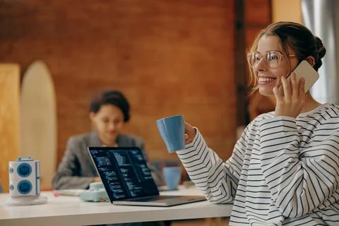 Young female computer programmer is talking phone while coding at laptop sitting Foto stock