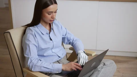 Young female freelancer with bionic hand is using laptop and sitting in chair at Stock Footage 195079868