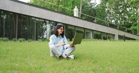Young female freelancer working remotely in a city park, sitting on the grass Stock Footage 317909678