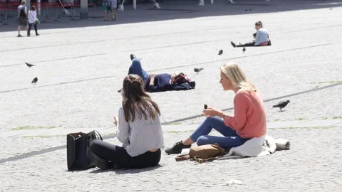 Young female friends sit on pedestrian square cobblestone eating and relaxing Stock Footage