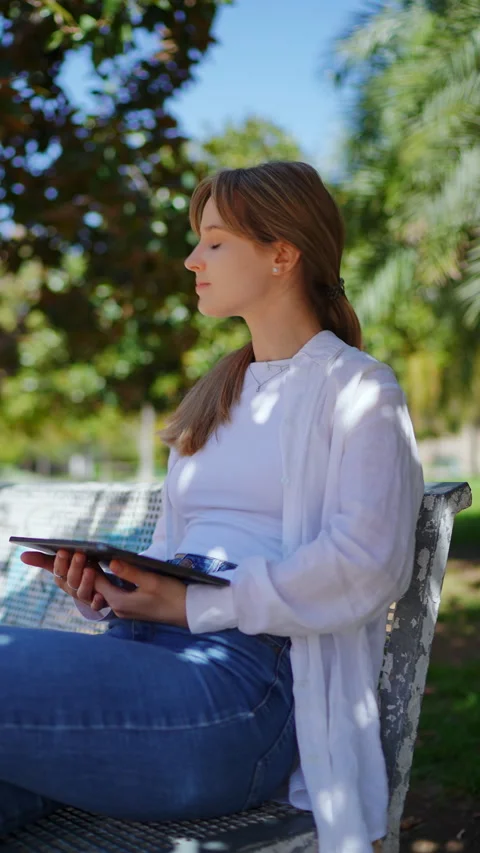 Young female interacting with digital tablet while resting on wooden park bench Stock Footage 308388364