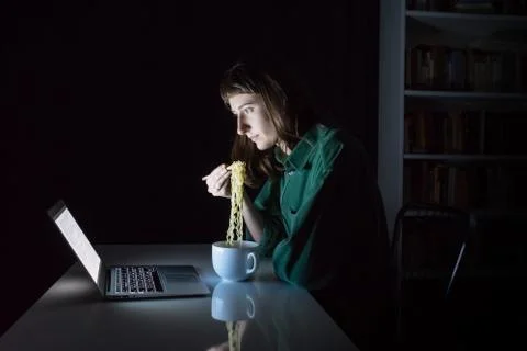 Young female at laptop computer eats instant ramen noodles with chopsticks Stock Photos