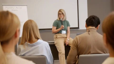 Young female lecturer talks using microphone, sitting on a table Stock Footage 218615799