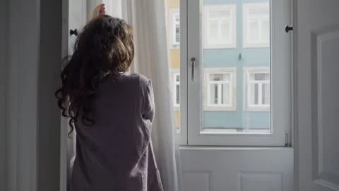 Young female opens apartment window curtain and smiles looking outside to street Stock Footage 293495950