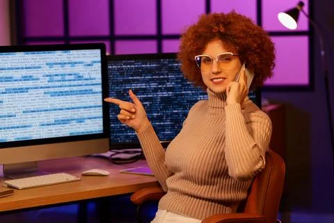 Young female programmer talking by phone and pointing at computer screen in o Foto stock