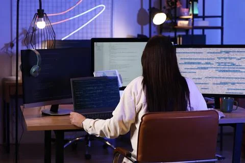 Young female programmer working with laptop in modern office at night, back v Stock Photos