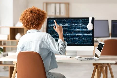 Young female programmer working with laptop and computer in office, back view Stock Photos