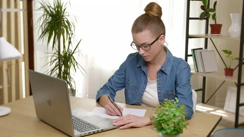 A young female student writes in a notebook lesson while looking into a laptop.  Stock Footage 201949513