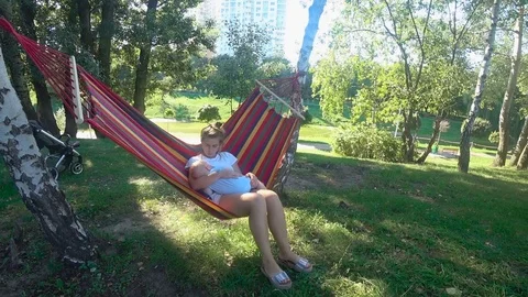 Young female swings in a hammock with baby son infant breast feeding Stock Footage