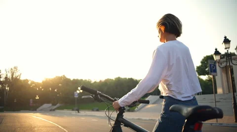 Young female talks on the phone while walking next to her bike in park Stock Footage 68346275