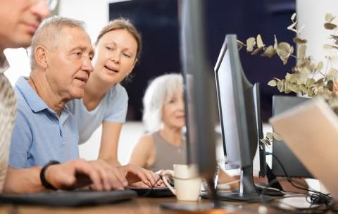 Young female teacher of computer class for seniors assisting man Stock Photos